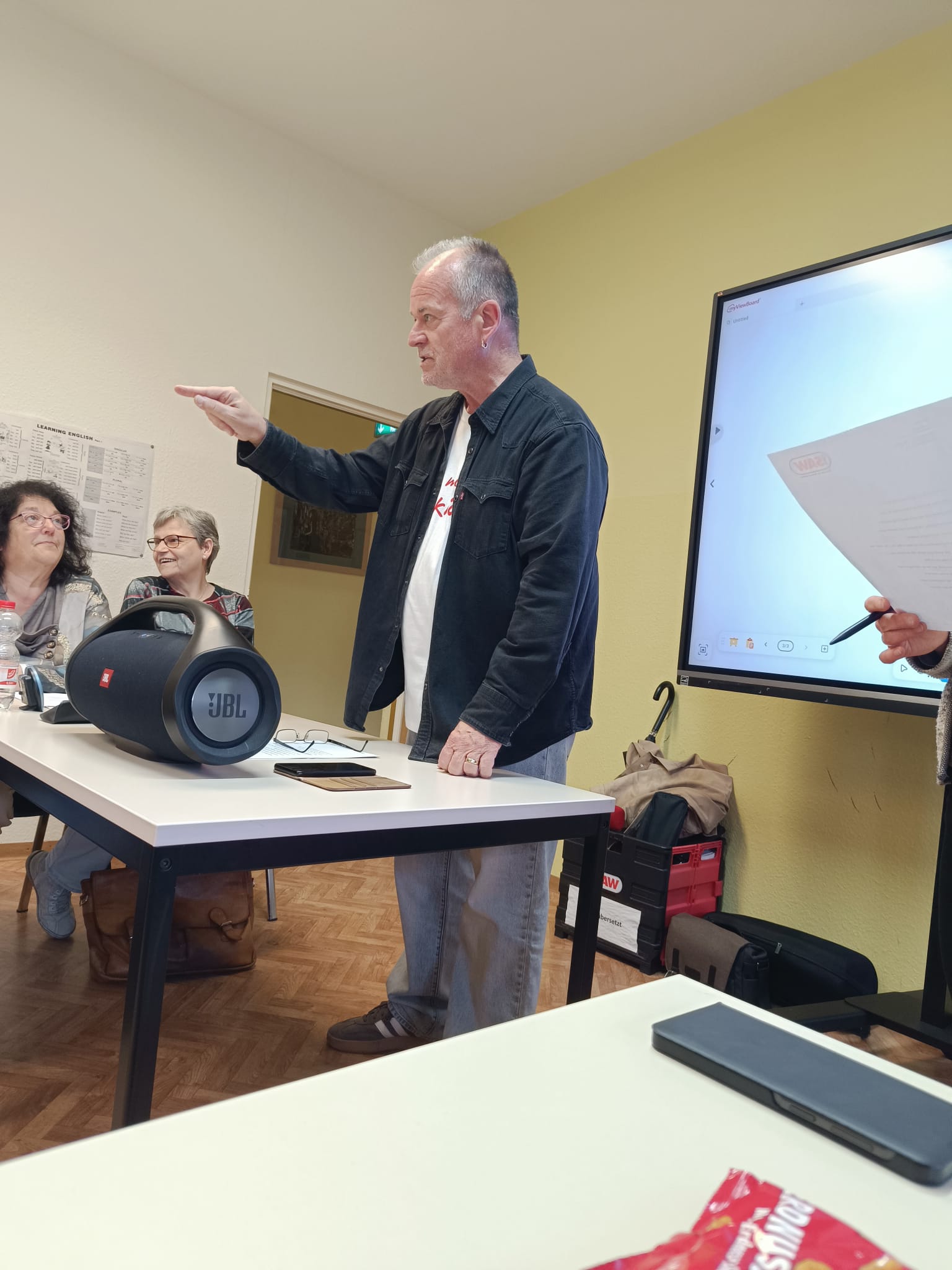Ein Mann hält einen Vortrag in einem Kursraum vor einem digitalen Whiteboard; im Vordergrund Kursteilnehmerinnen und eine JBL-Box auf dem Tisch.
